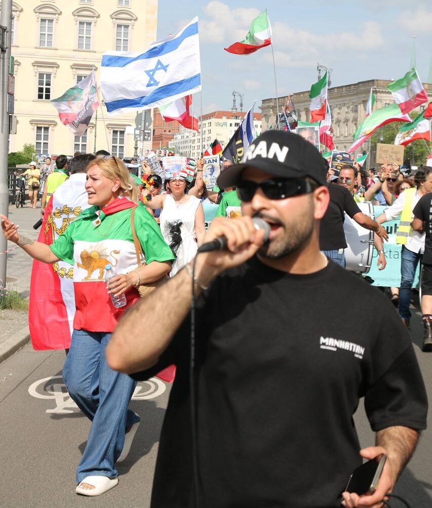 People at a rally hold Israeli and Iranian flags while a speaker talks into a microphone.