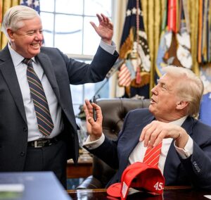 Donald Trump seated at the Resolute Desk with Lindsey Graham standing beside him in the Oval Office.
