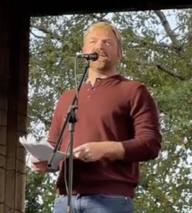 Graham Platner speaking at a microphone during an outdoor event in Brunswick, Maine.