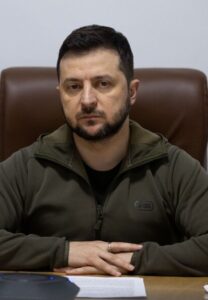 Ukrainian President Volodymyr Zelenskyy sits at a desk beside a Ukrainian flag.