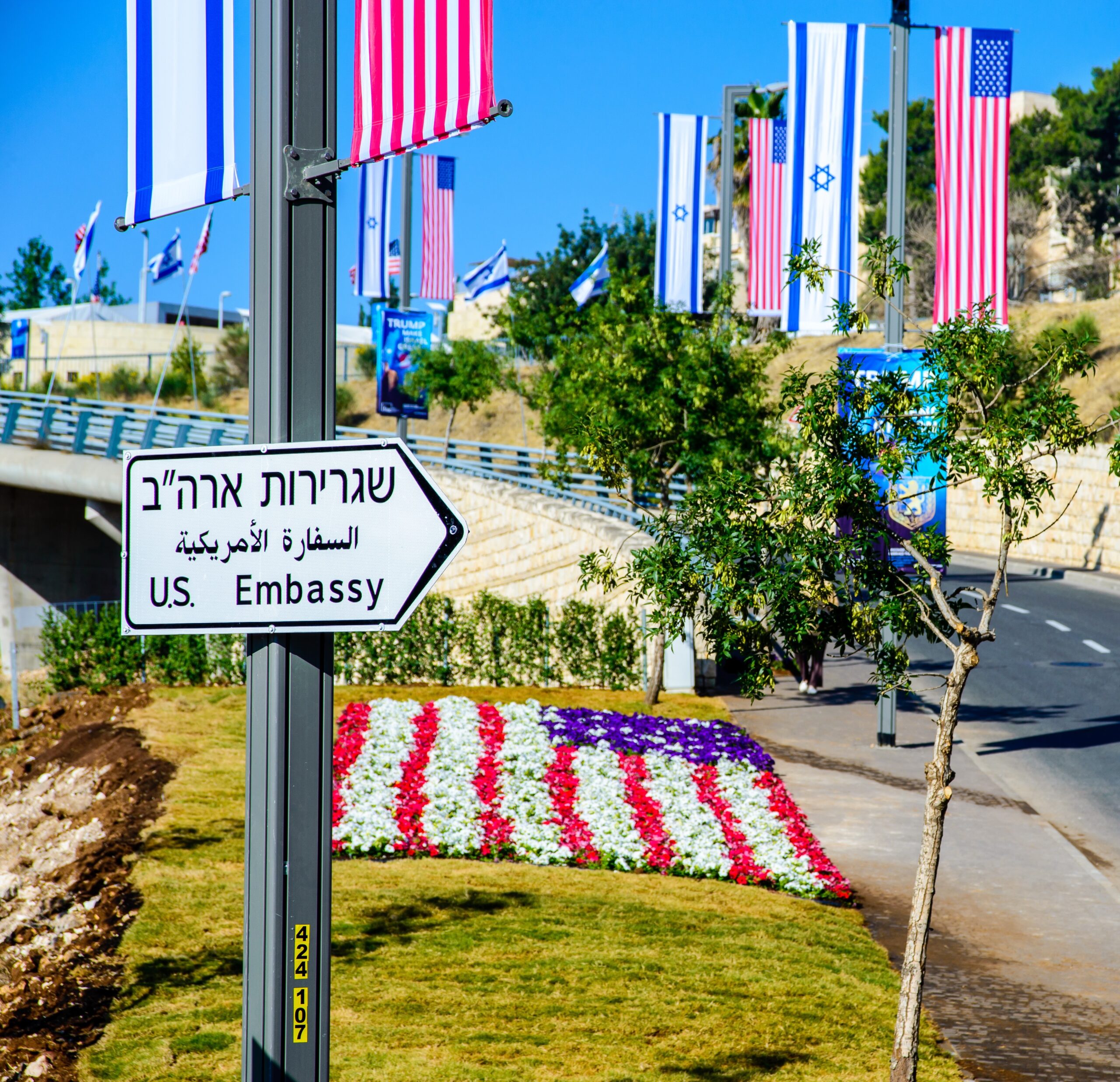 A sign pointing to the U.S. Embassy (in English, Hebrew, and Arabic) in Jerusalem with American and Israeli flags displayed along the road, while a bed of red, white, and blue flowers makes up the American flag in the grass.