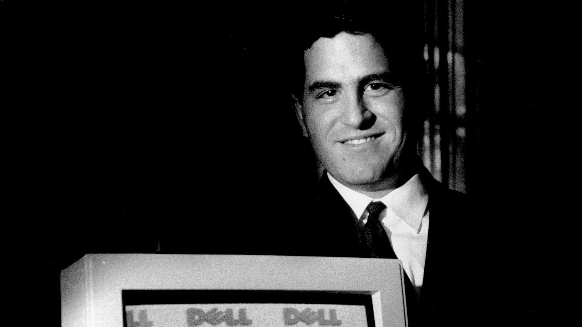 A young Michael Dell smiles while standing behind an early Dell computer.