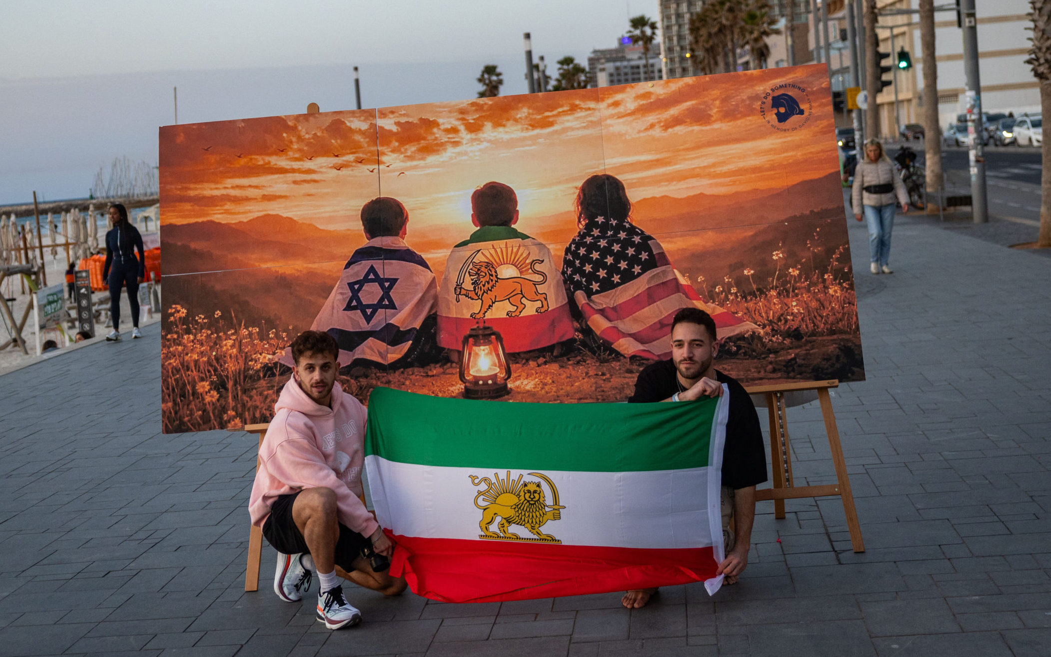 Two people holding the Iranian opposition flag in front of artwork showing Israeli, American and Iranian opposition symbols in Tel Aviv.
