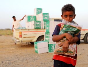 Displaced Yemeni child holding a bag of food aid beside relief supplies in Hajjah Governorate.