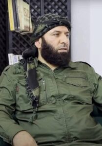 A man in military-style clothing sits on a sofa indoors.