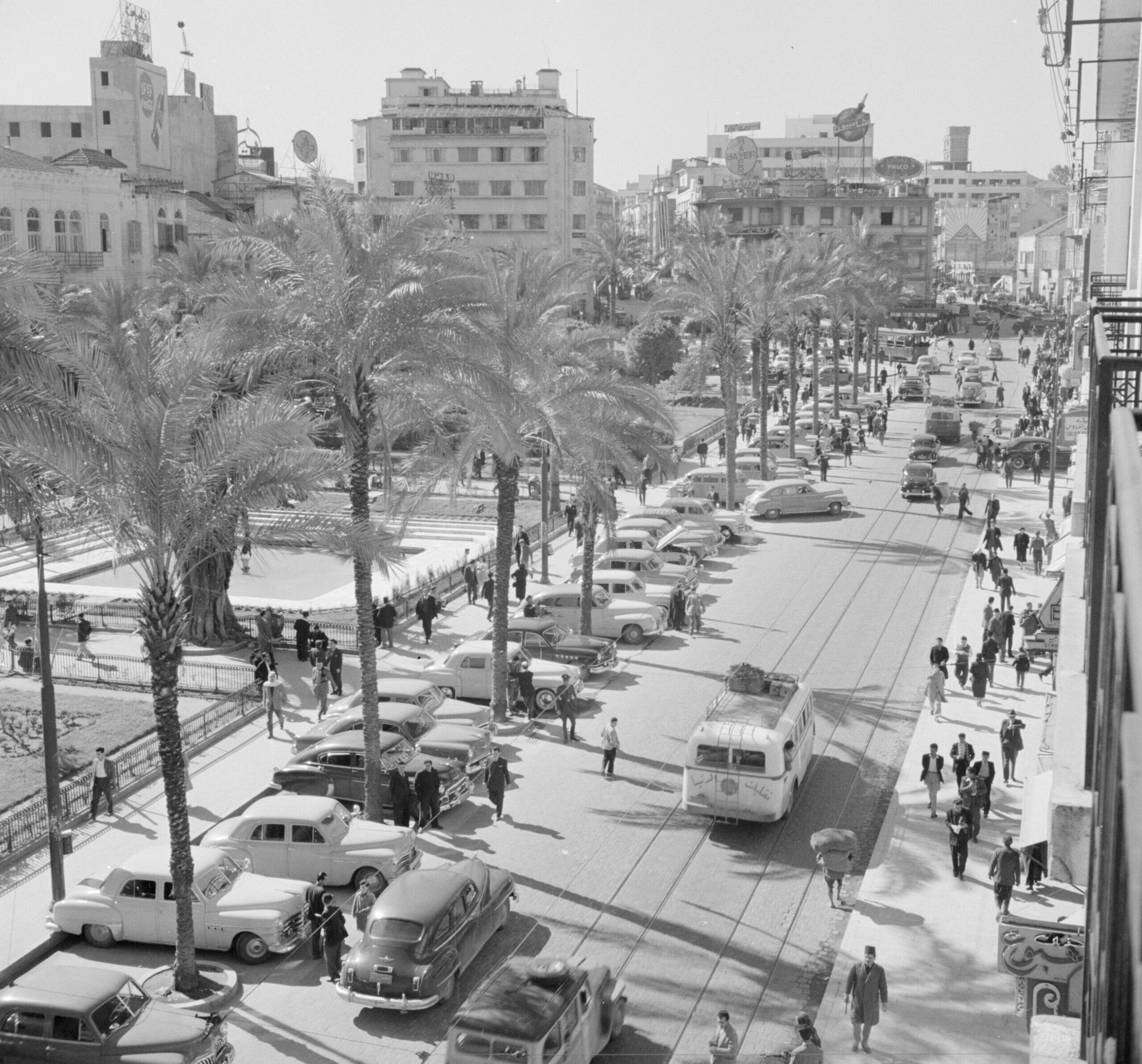 Historic black-and-white street scene in Beirut with palm trees, cars, pedestrians, and city buildings.