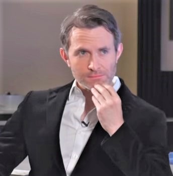 Author and journalist Douglas Murray looking pensive in a black suit jacket against a neutral studio background
