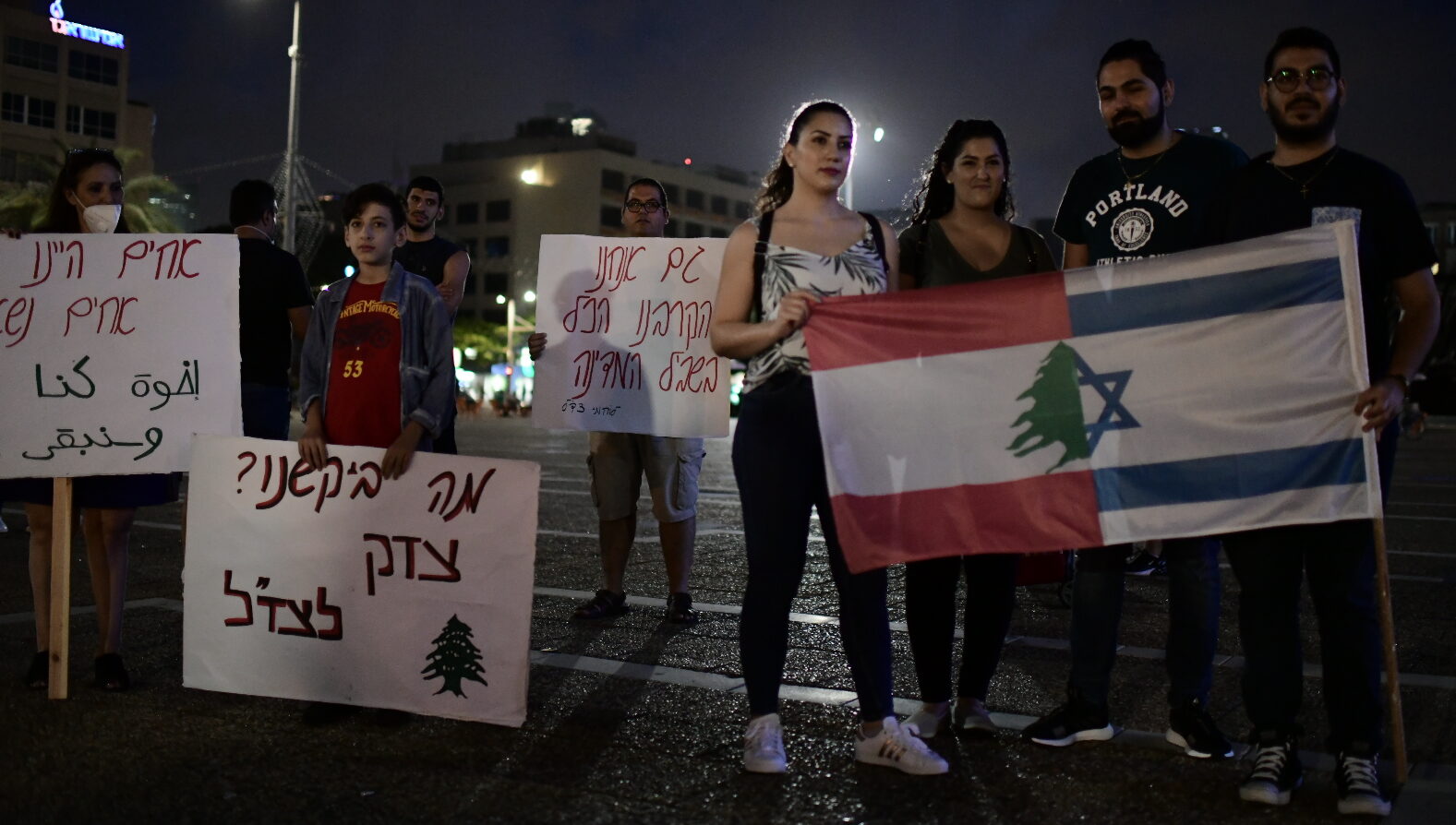 People stand at a rally in Tel Aviv holding an Israeli-Lebanese flag and signs calling for peace.
