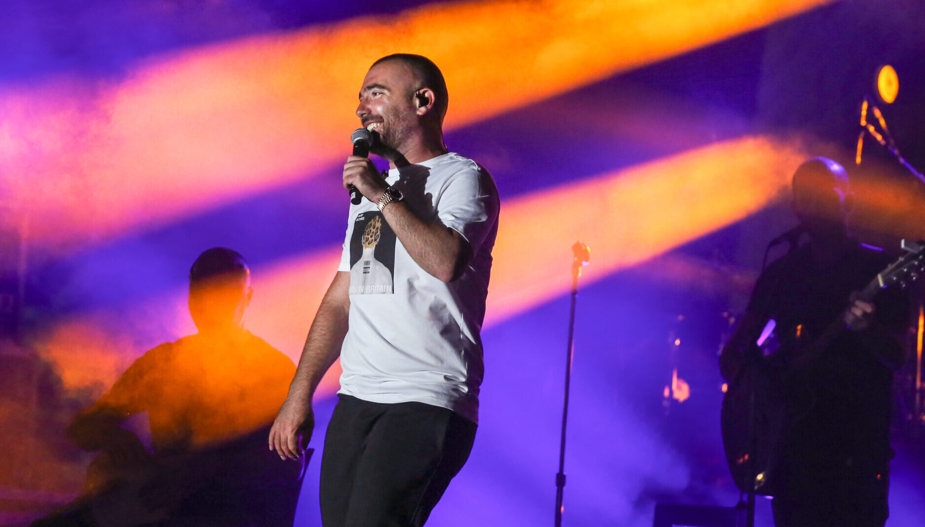 Omer Adam performs on stage with colorful lights behind him.