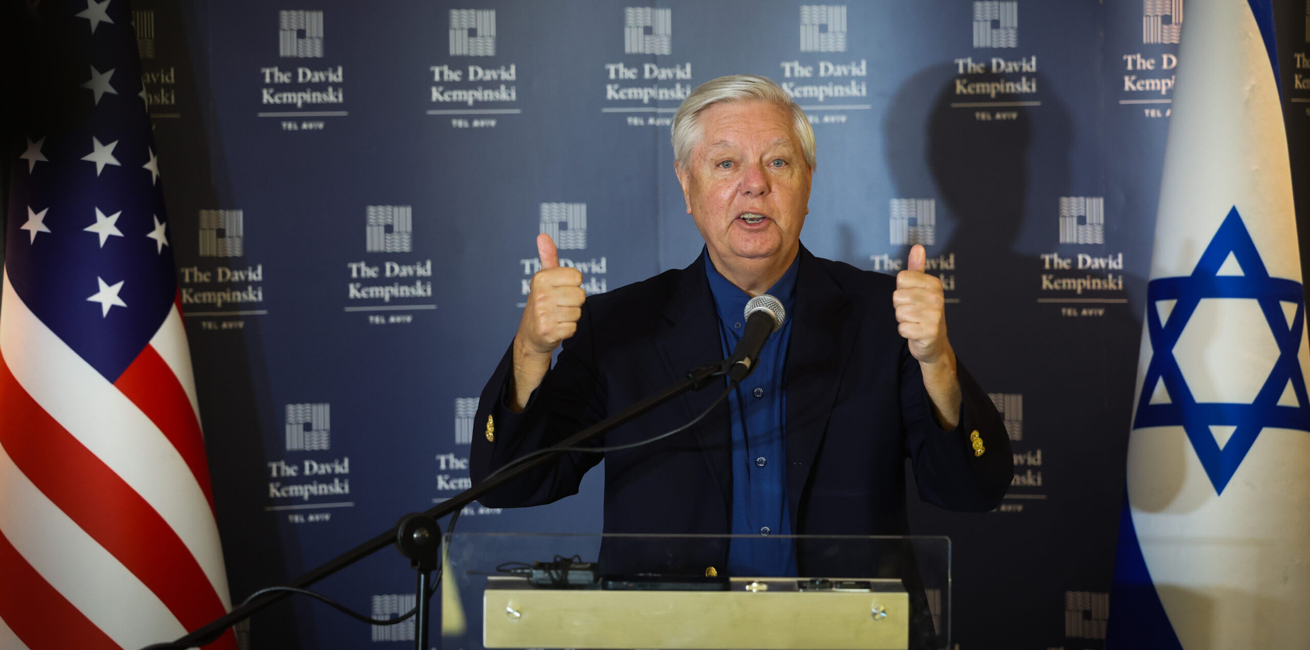 U.S. Senator Lindsey Graham speaks at a podium in Tel Aviv with Israeli and American flags beside him.