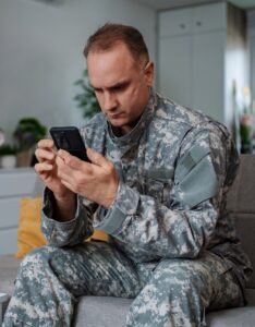 A soldier in uniform looking at a smartphone while sitting indoors.