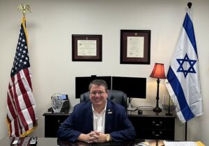 Randy Fine seated at a desk in his office with American, Israeli, and Florida flags behind him.