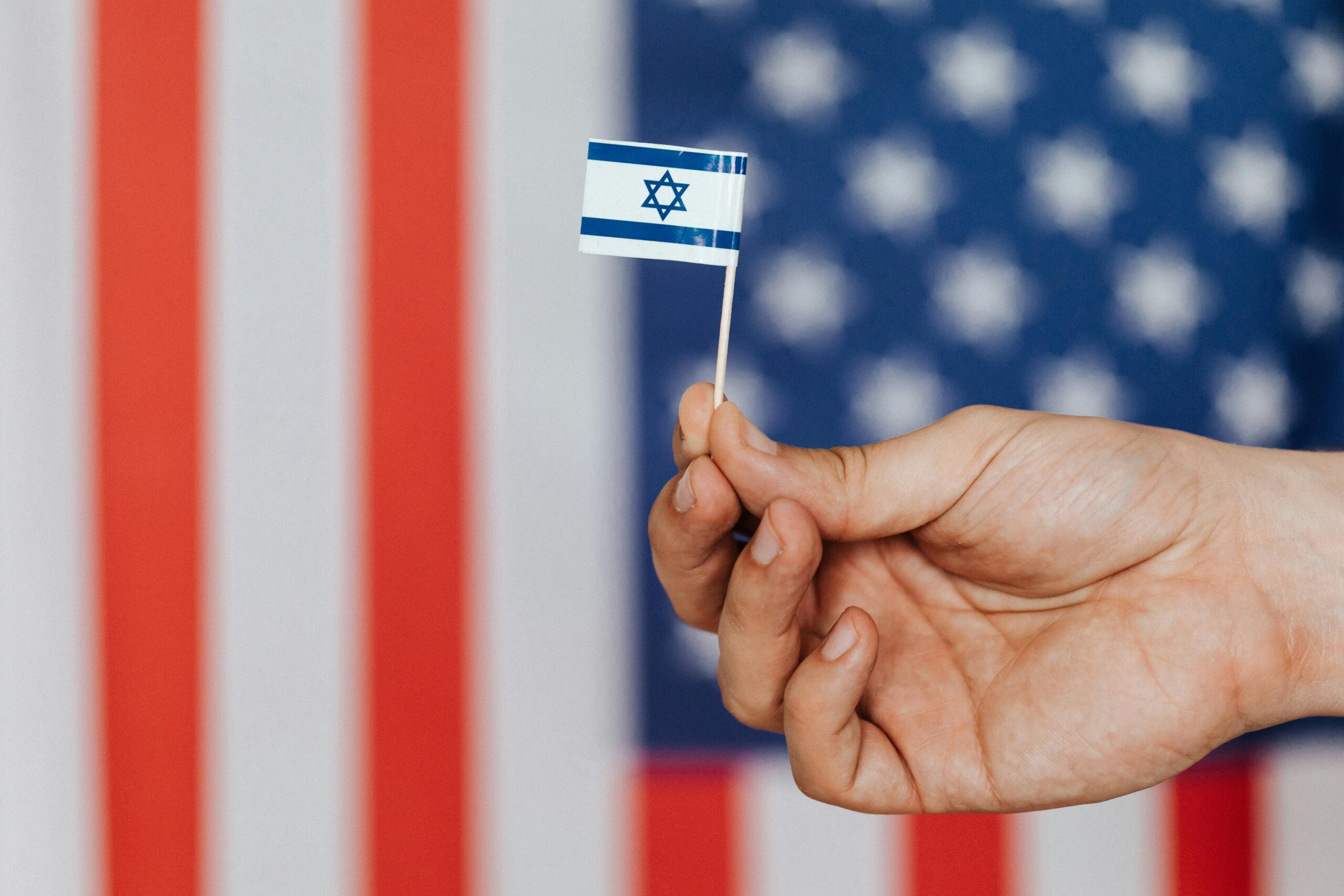 Hand holding Israeli flag, with backdrop of American flag