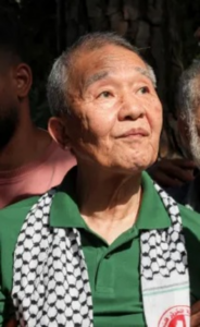 Kōzō Okamoto wearing a green shirt and a keffiyeh-style scarf.